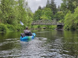 man in a blue sit-in kayak on the Black River. A bridge over the river can be seen in the distance